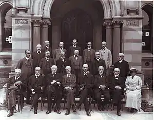 A group of people pose for a formal group portrait beneath a stone arch. There is only one woman in the group. While the men have removed their hats, she still wears hers.