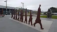 an iron oxide metal sculpture depicting abstract figure walking in single file, a stone sculpture in the background of a mother and two children, the entrance of the single story railway station building on the left