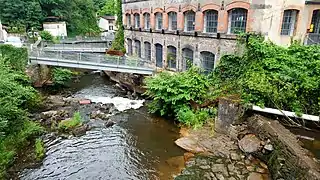 Overhead view of the May factory. The left half is taken up by the Durolle River, crossed in this section by 3 metal bridges. The right half shows the factory facade with 2 floors of workshops, each with 8 large arched windows. On the lower level, the windows are arched with cut stone, while on the upper level they are arched with red brick.