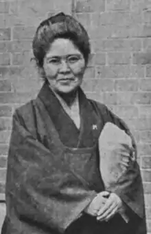 A middle-aged Japanese woman, standing in front of a brick wall. She is wearing a simple, dark, kimono jacket, holding a bundle, hands clasped. She wears glasses and her hair is dressed in an updo.