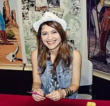 Portrait photo of a seated young woman with wavy brown hair who is wearing a white hat and blue jeans vest, and is smiling at the camera, while holding a pink pen.