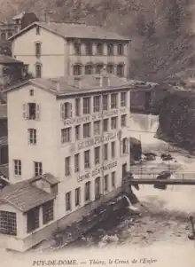 Antique postcard in bistre and white showing the center of the valley with the Durolle just in front of the waterfall and, on the left, the Entraygues factory buildings rising in a 4-story parallelepiped with 7 rectangular windows per level. The masonry belts between the floors read clearly, from top to bottom: Manufacture de Coutellerie Fine, Sauzede Père et Fils, Usine d'Entraygues.