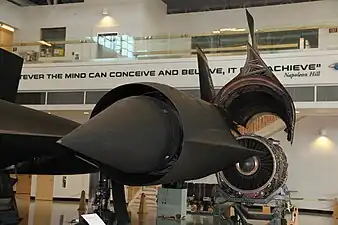 A view of the entry to an SR71 Mach 3.2 mixed external-internal inlet looking in direction of airflow to the engine. The centre translating cone has 26 inches of travel between extended, up to M1.6 (shown), and fully retracted at M3.2. An oblique shock from the cone tip, an internal oblique shock from the cowl lip and a normal shock give the required pressure recovery at M3.2. The boundary layers on the cone and cowl inner surface have to be removed before the final shock-wave where the flow becomes subsonic. Otherwise shock-induced separation occurs. The two removal features are just visible. The cone boundary layer is removed through the band of holes (porous bleed). The boundary layer on the cowl inner surface is removed through a shock-trap bleed. This ram bleed is just visible on the lower surface in front of a row of streamlined lumps called "mice" which reduce the rate of diffusion.