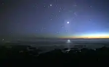 A photograph of the night sky taken from the seashore. A glimmer of sunlight is on the horizon. There are many stars visible. Venus is at the centre, much brighter than any of the stars, and its light can be seen reflected in the ocean.