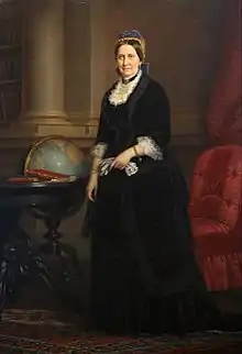 Portrait of a standing woman wearing a blue cap, black dress with white lace at the neck and cuffs, and holding a pair of gloves. To her right is a globe and to her left is an arm-less upholstered chair.