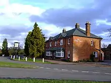 A substantial brick building at a bend in the road, with pub sign reading "Verney Arms"