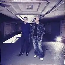 The band members standing in an industrial space, one looking toward the camera and the other to the side