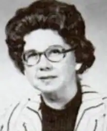 A middle-aged white woman with dark hair in a bouffant style, wearing glasses and a striped jacket over a dark top