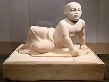 Beige marble statue of a stout young child aged about two years old lying on his left side. The child's head is shaven, his eyes gaze over the viewer's shoulder and his lower body is covered in a draping cloth that hangs limply between his flexed feet. The child supports his torso with his left hand in which he holds an unidentifiable object, he also holds a small bird in his right hand. The sculpture rests on a heavy socle inscribed with barely visible letters spanning the upper part of the socle vertically.