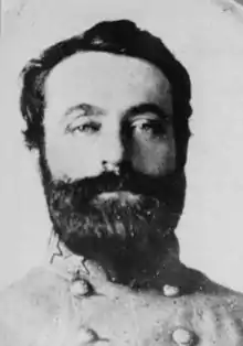 Black and white photo shows bearded, dark-haired man staring straight into the camera. His gray military uniform has stars on the collar.