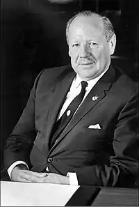 Half-length portrait of mustachioed man in suit and tie, seated at a desk