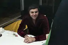 A Caucasian man wearing a dark burgundy shirt sits at a table.