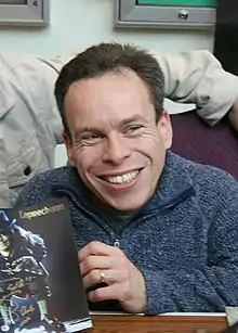 Actor Warwick Davis sits at a table, smiling and holding Leprechaun merchandise.