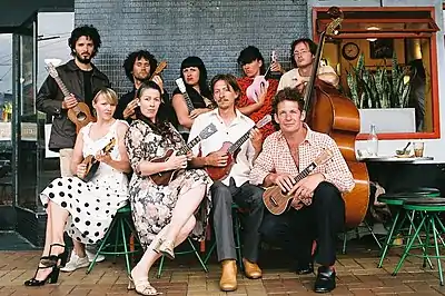 group of musicians with their instruments some sitting some standing with pensive and happy expressions in front of a window and tiled cafe building