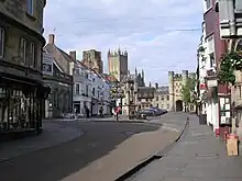 Street scene with shops on either side of the street. In the centre stands an old stone monument. In the background are an old stone gatehouse and behind it the towers of the cathedral.