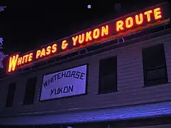 A red electric sign is seen at night, displaying words "White Pass & Yukon Route" above a white, wooden sign emblazoned with "Whitehorse Yukon".