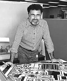 A man with a beard leans over a table containing technical drawings of an electronic circuit