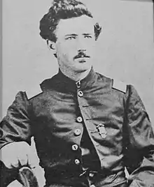 Head and torso of a white man sitting sideways in a chair, his right arm resting on the chairback. He has a thin mustache, curly hair, and is wearing a military jacket.