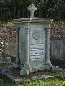 A granite, squared plinth with a column at each corner and a small cross on top of it, among other headstones