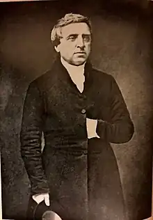 Black and white photograph of Matthews holding a hat by his side in his right hand and with his left hand tucked in between his coat buttons