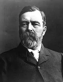 A black and white portrait photo of a thickly bearded man in a wool coat.