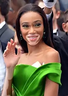 The image features a woman (Winnie Harlow Cannes) with a smiling expression, waving her hand. She has vitiligo, characterized by patches of skin with depigmentation. Her hair is styled straight and parted to the side. She is wearing makeup with prominent eyelashes and eye shadow. She is dressed in an off-the-shoulder, vibrant green garment. Earrings are visible, and the setting is an event with other people in the background.