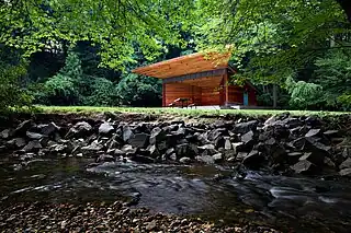 A small, boxy, wooden stage with a trapezoidal overhang stands in the center of meadow. In the foreground is a running stream with a stone embankment.