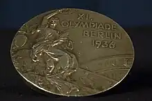 A tarnished gold medal featuring a person in a toga, and the words "XI Olympiade Berlin 1936".