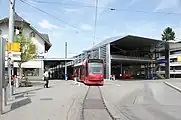 tram of line 6 between the station building (left) and the train shed of the Worb Dorf-Worlaufen railway (right) engaging in the track loop in the foreground, with a bus stop in its center and hiking fingerpost to the left (2012)