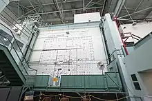 Two workmen on a movable platform similar to that used by window washers, in front of a wall with arrays of holes and many wires running across it. A sign says "Graphite Reactor loading face".