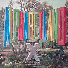 A painting of a colorful logo with tall letters reading "ALPHABETLAND" in a forest with a lake that has a grey "X" below it on the ground