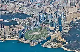 An image of Xinghai square and the surrounding areas. A large open green space is seen amid a sprawling urban city center and adjacent ocean.