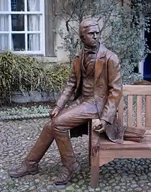 Bronze statue of Darwin in 1830 clothes, seated on the arm of a wooden bench;  behind him plants partly cover a stone wall, a window has white-painted wooden frames