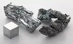 A crystal of a silvery-colored metal, a crystal of a dark metal and a cube of metal standing on a light grey surface