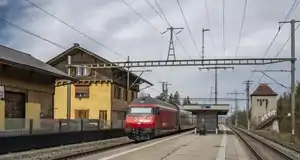 Red locomotive leads train past platform; there is three-story building with gabled roof at left